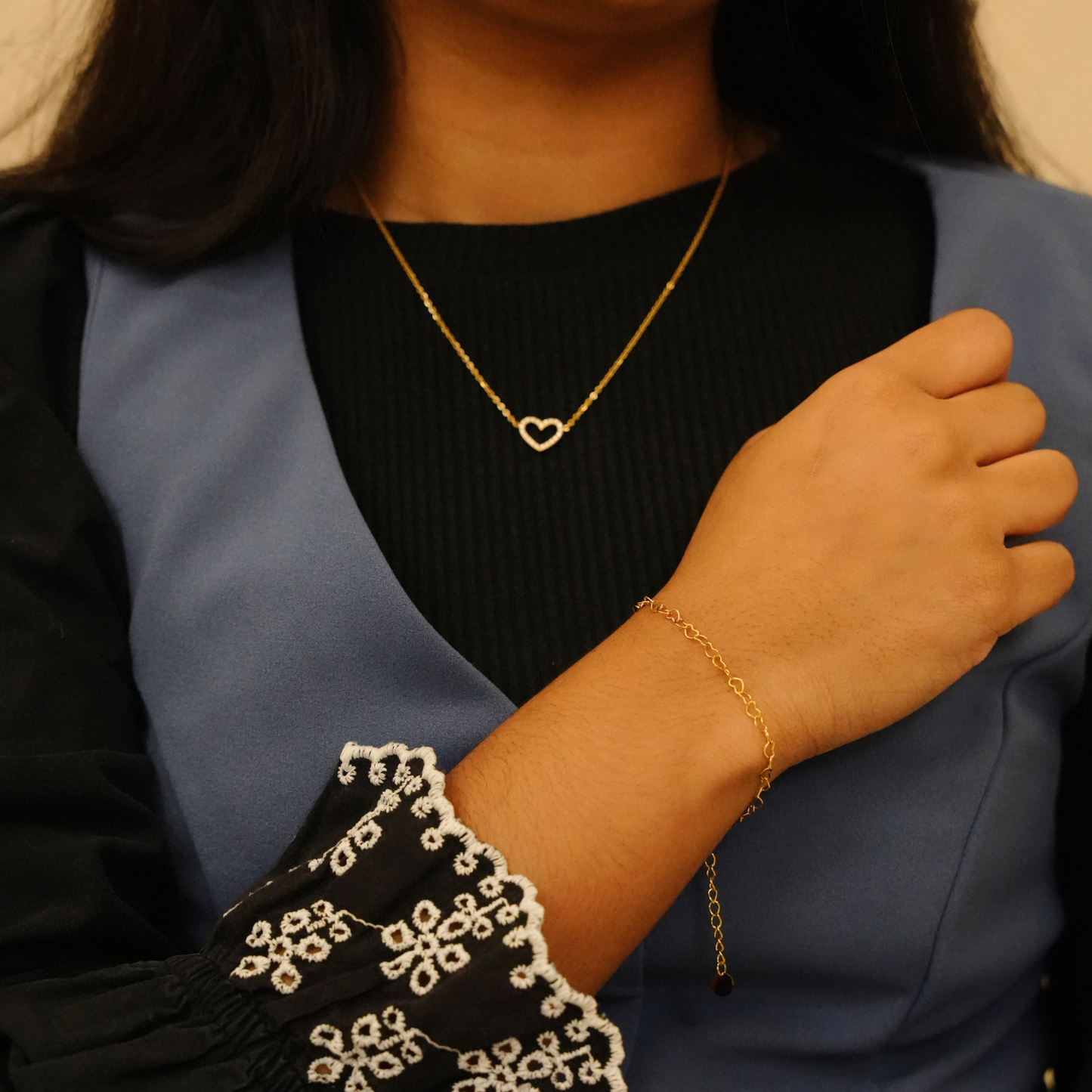 Gold heart pendant necklace with crystal accents, on a delicate chain, worn with a gold bracelet and a blue vest.
