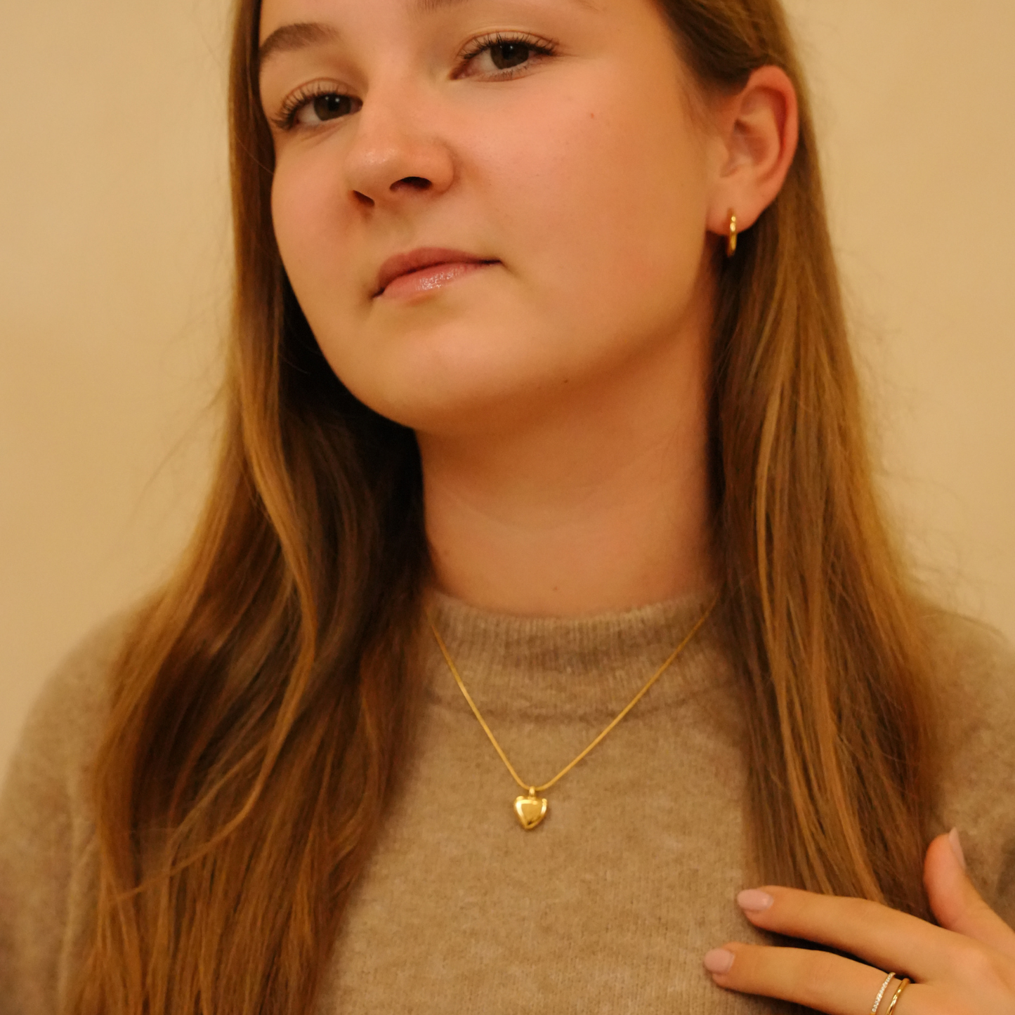 Gold heart pendant necklace on a delicate chain, featuring a heart-shaped locket, styled with a tan sweater and gold hoop earrings.