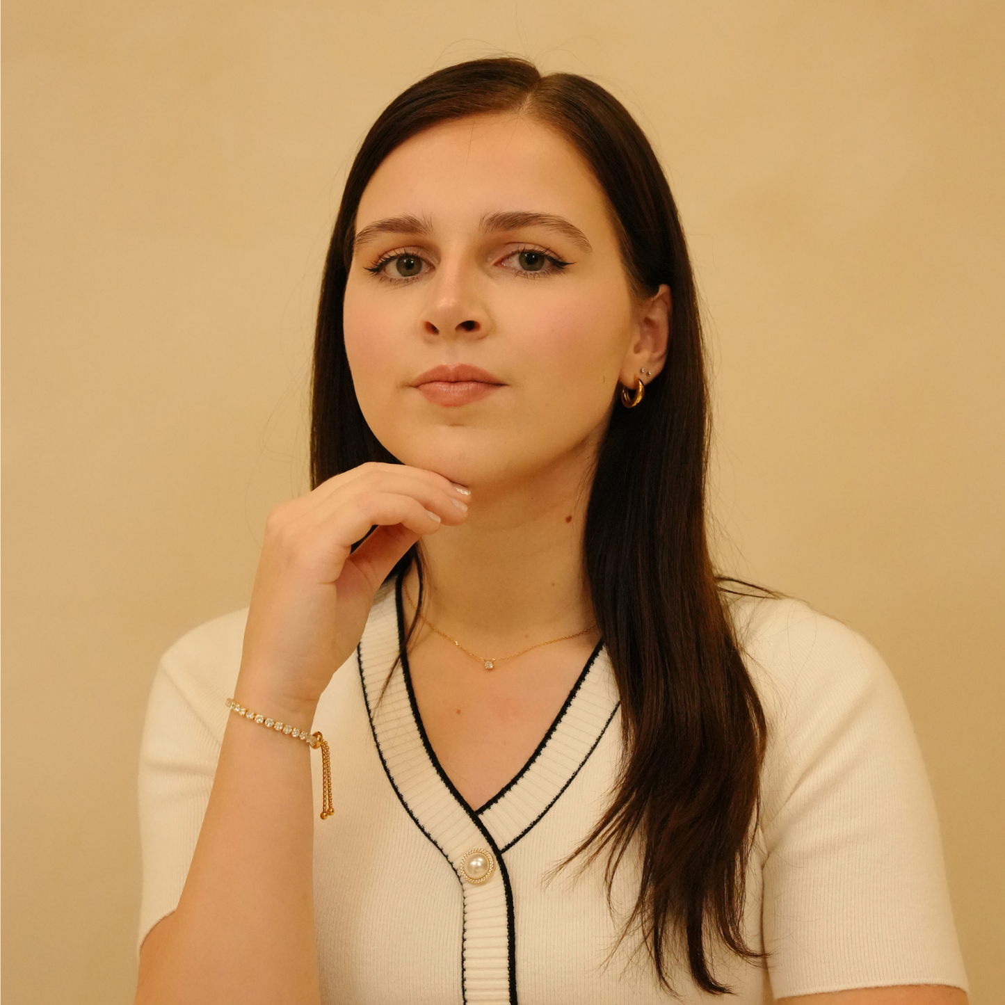 Gold-toned bracelet by Julia featuring small stones and a dangling chain, worn with a white cardigan and gold hoop earrings.