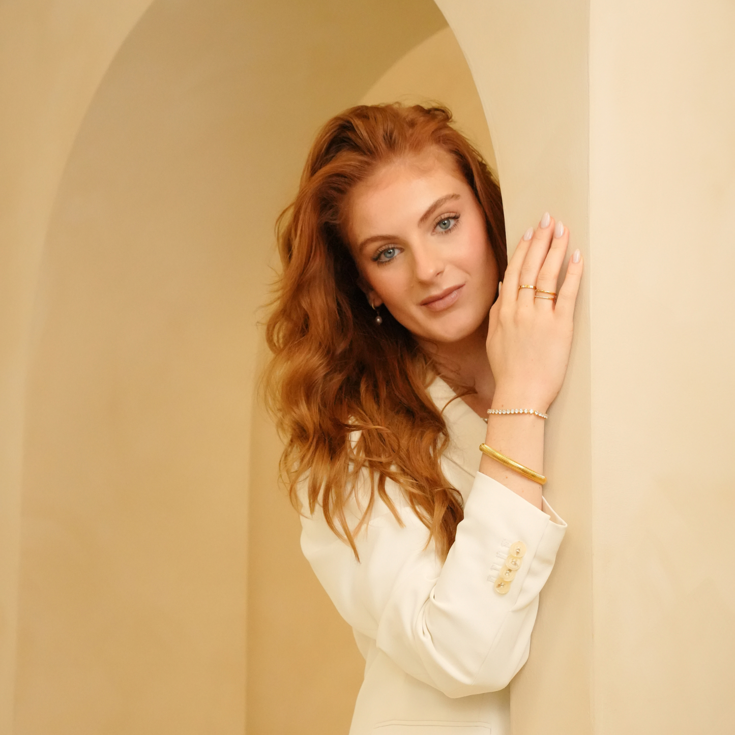 Gold flexible bracelet by unknown brand, worn with a white blazer and rings, resting against an archway.