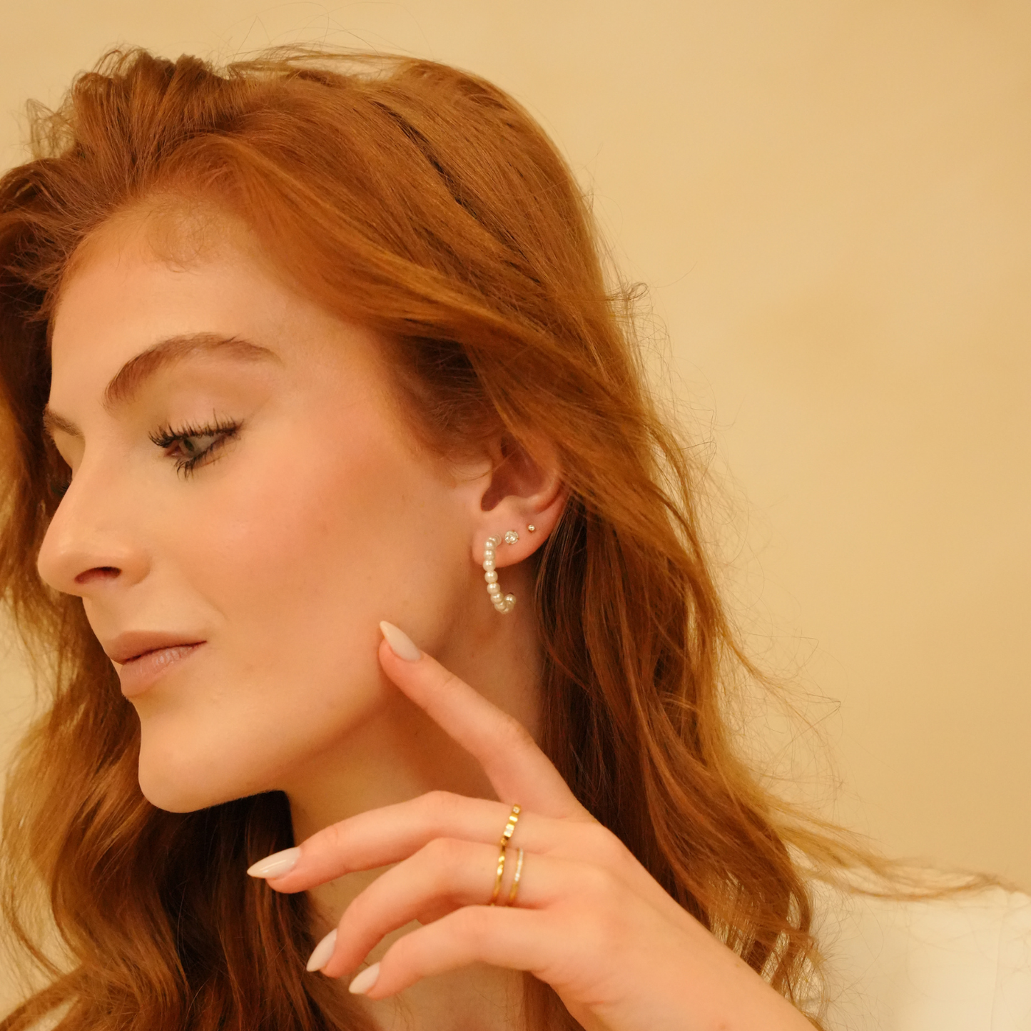 Pearl hoop earrings by an unknown brand, shown on a model with red hair, with gold rings on the fingers.