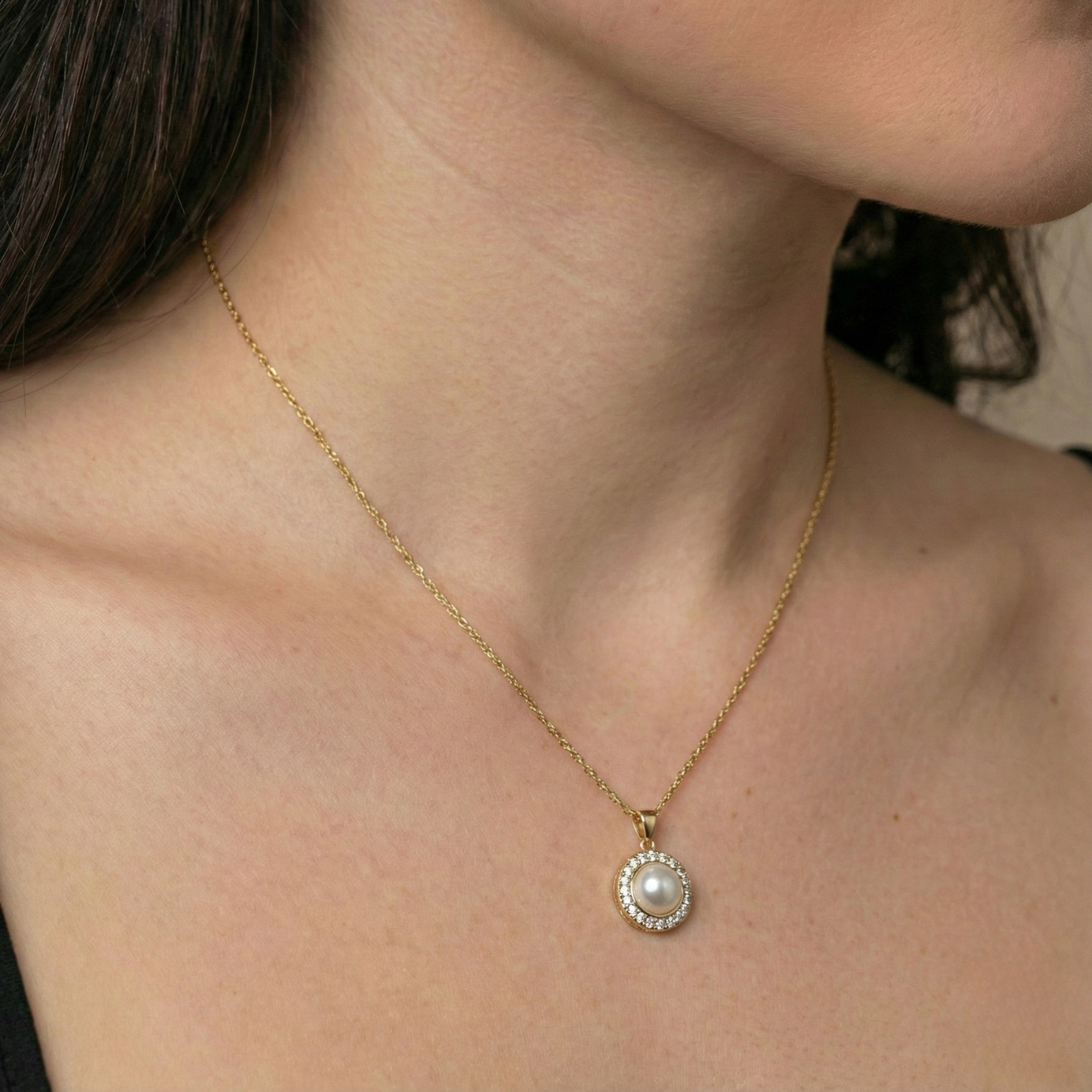 Gold pearl necklace by Amour, featuring a round pendant with crystal halo, worn on a woman.