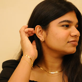 Gold zirconia earrings with hoop and stud accents, positioned near the ear, and styled with a gold herringbone necklace and bracelet.
