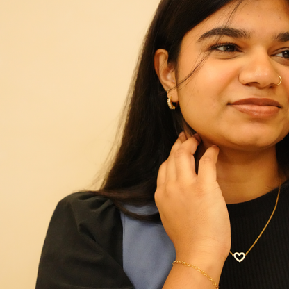 Heart pendant necklace with gold chain, worn with gold hoop earrings and a gold bracelet.