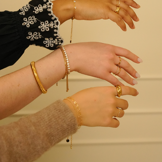 Gold wavy zirconia ring with stone detailing, worn on a hand with multiple gold rings and bracelets.
